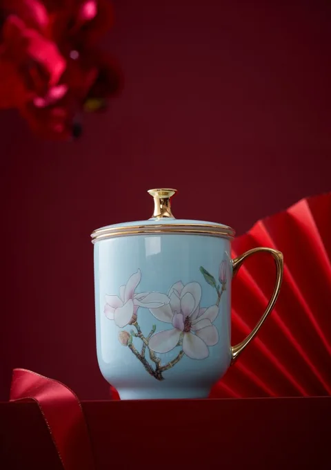 A blue ceramic mug with floral decoration and a gold-coloured lid, styled against a red background.
