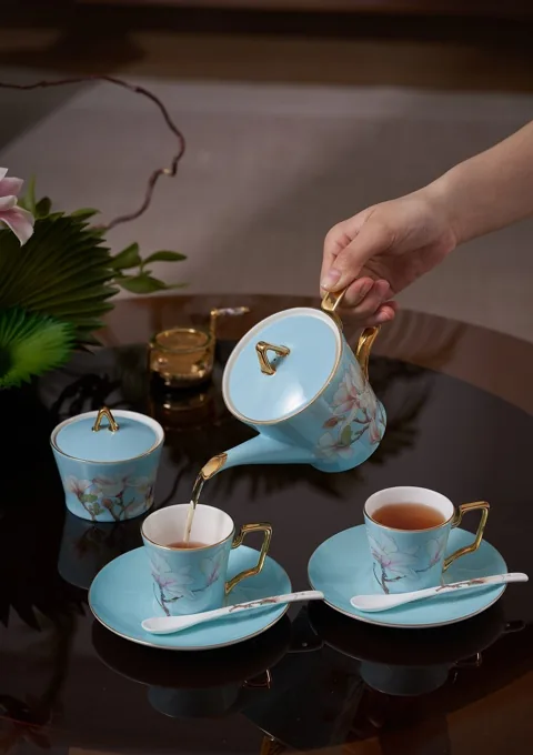 A coordinated floral ceramic tea service shown during a tea-pouring moment in a dark interior.
