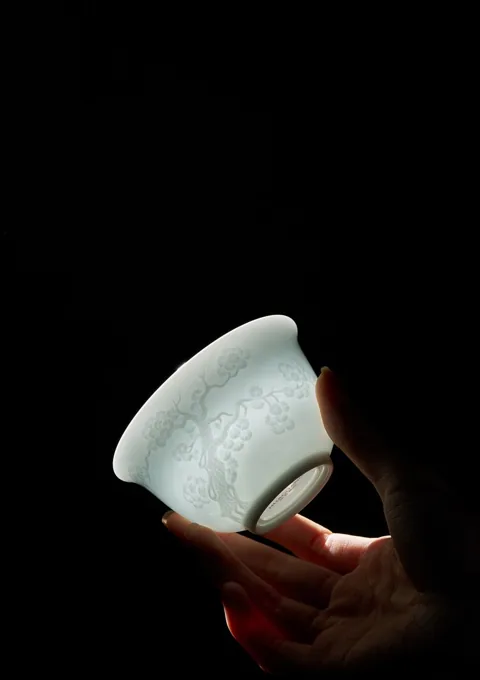 A hand holding a small translucent ceramic cup against a dark background.