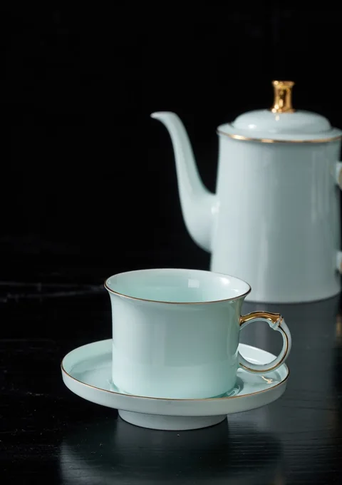 A pale blue ceramic coffee pot, cup and saucer styled against a dark background.