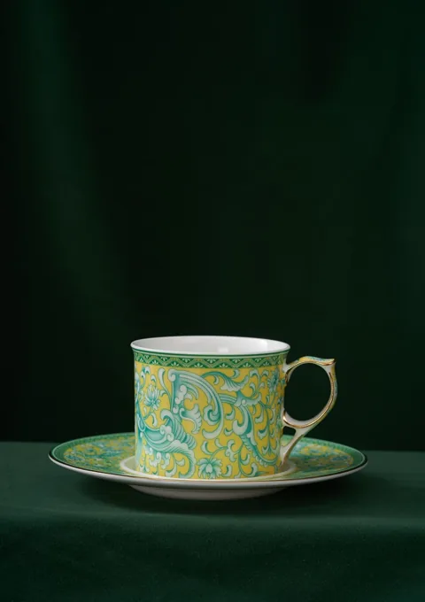A green floral ceramic cup and saucer styled against a dark green background.