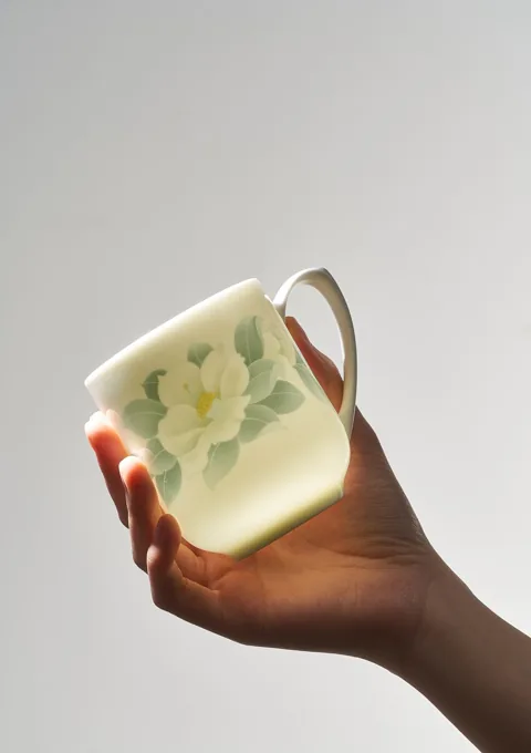 A hand holding a pale ceramic mug with a floral motif, illuminated from behind.