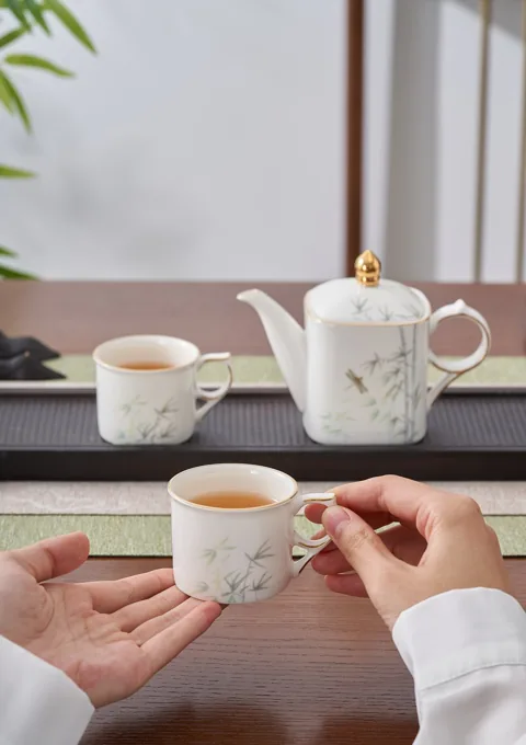 Hands holding decorated ceramic cups beside a coordinated teapot in a relaxed tea setting.