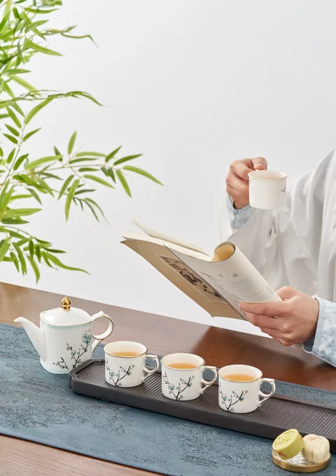 A ceramic tea service arranged beside a seated person reading in soft daylight.