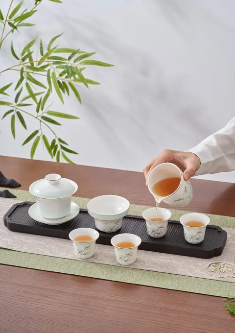 A hand pouring tea into a small ceramic cup beside a coordinated lidded bowl and tea tray.