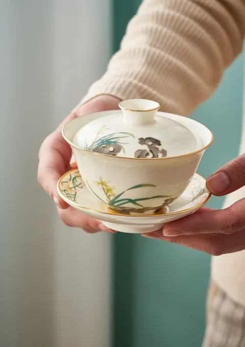 Hands presenting a decorated ceramic lidded tea bowl and matching saucer.
