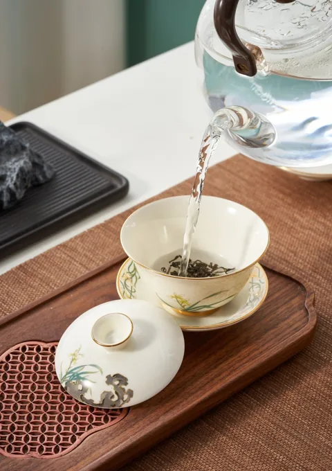 Water poured into a decorated ceramic tea bowl on a tray beside a matching lidded cup.