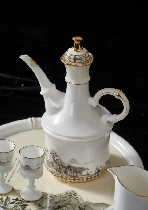 An ornate white ceramic pouring vessel with gold-coloured detailing and a matching cup.