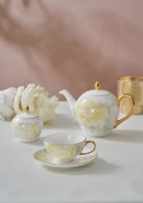 A light floral ceramic teapot, cup and saucer styled with pale flowers against a warm neutral background.