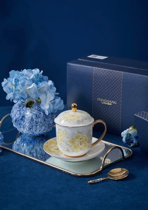 A decorated ceramic mug with a lid beside blue gift packaging and hydrangeas.