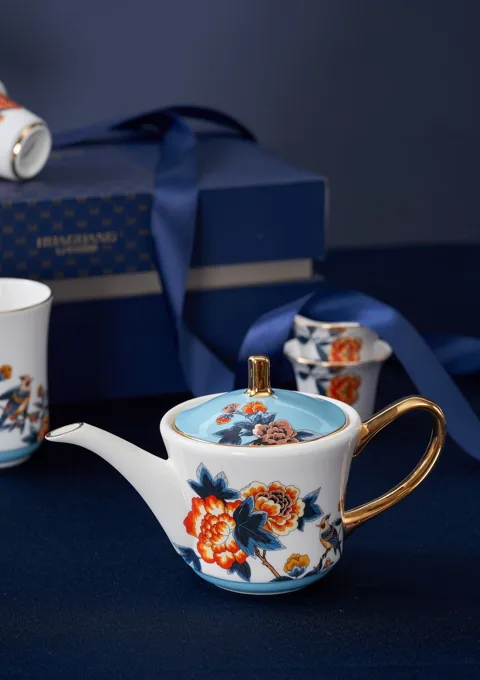 A colourful floral ceramic teapot displayed beside blue presentation packaging.