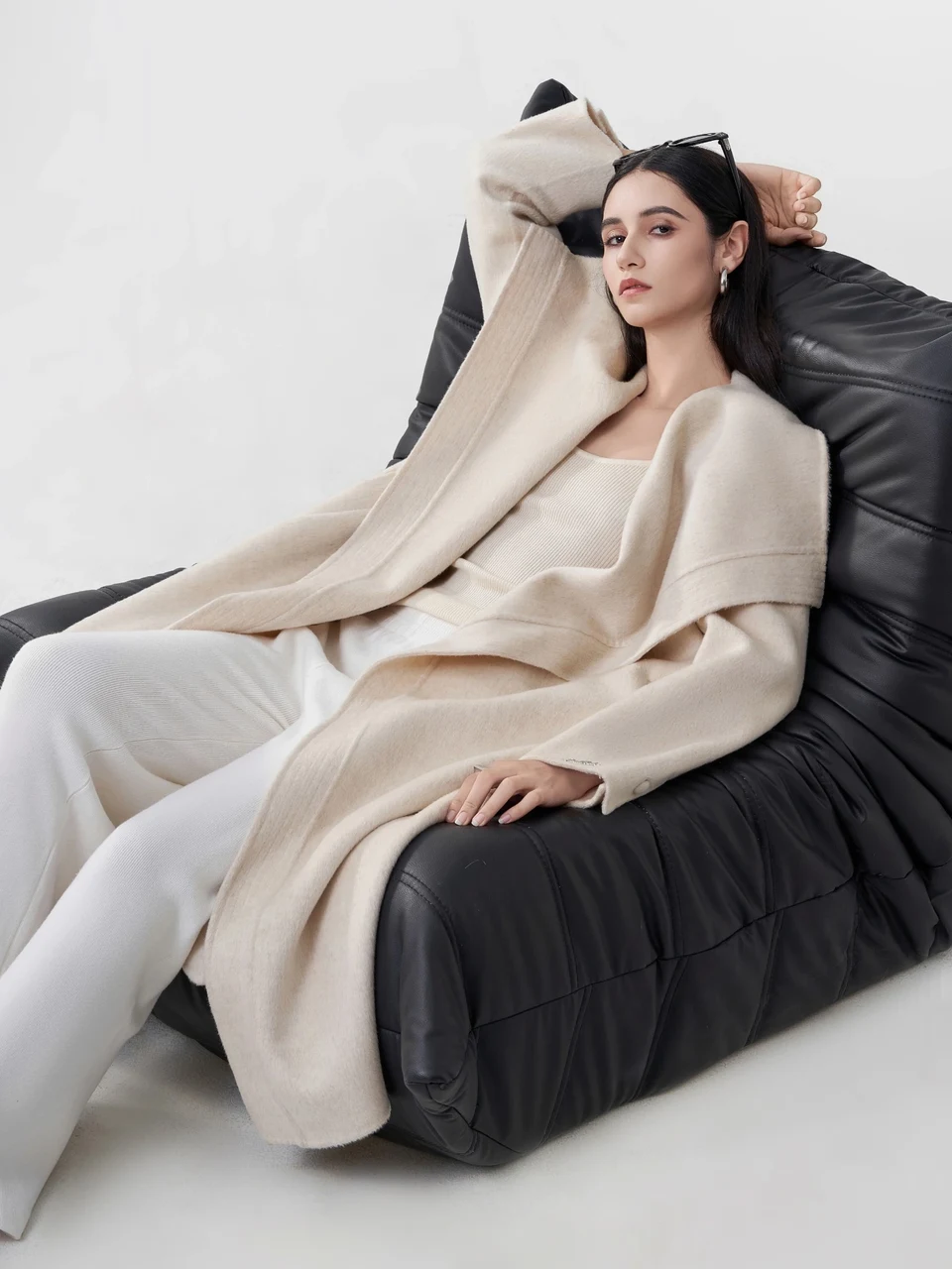 A model in a cream coat reclining on a black lounge chair.